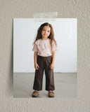 Młoda dziewczyna z długimi brązowymi włosami stoi w pomieszczeniu, ubrana w jasnoróżową koszulę w kwiatowe wzory, luźne brązowe spodnie i buty w stylu mokasynów. Zdjęcie jest przyklejone taśmą do beżowej ściany.