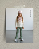 Młoda dziewczyna stoi w pomieszczeniu ubrana w różową czapkę i szalik, biały sweter, zielone rozkloszowane spodnie i białe trampki. Jest zwrócona do przodu z neutralnym wyrazem twarzy, na prostym tle.