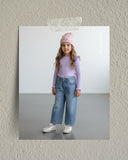 Młoda dziewczyna stoi w pomieszczeniu, ubrana w liliową bluzkę z długim rękawem, luźne niebieskie dżinsy, białe trampki i różową czapkę. Ma długie falowane włosy i uśmiecha się delikatnie do aparatu. Zdjęcie jest przyklejone taśmą do teksturowanej beżowej ściany.