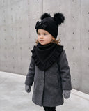 Dziewczynka w komplecie „Waffle” – czapka z pomponami i chusta z falbanką – w kolorze Czarny, przy betonowej ścianie.