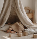 Dziecko leży pod beżowym kocem w materiałowym namiocie, z widocznymi tylko bosymi stopami. Drewniane klocki są rozrzucone na dywanie w przytulnym, delikatnie oświetlonym pokoju.