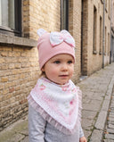 Dziewczynka w komplecie Flora w kolorze Jasny puder na brukowanej uliczce, zbliżenie na czapkę z uszkami misia i kokardą.