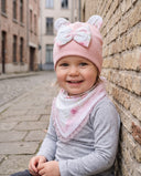 Uśmiechnięta dziewczynka w komplecie Flora w kolorze Jasny puder siedzi przy ceglanym murze.
