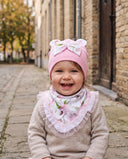 Roześmiana dziewczynka w komplecie Flora w kolorze Baby pink siedzi na brukowanej uliczce.