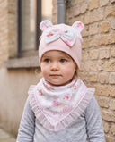 Dziewczynka w komplecie Flora w kolorze Łosoś przy ceglanym murze, zbliżenie na czapkę z uszkami misia i chustę z falbanką.