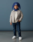 Małe dziecko stoi pod niebieską ścianą, ubrane w PanPan 35 Zestaw czapka z uszami królika i komin - bawełna, prążek, wraz z beżową pikowaną kurtką, niebieskimi dżinsami, białymi trampkami i jedną ręką w kieszeni.