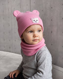 Dziewczynka w czapce z uszami misia i kominie w kolorze Baby pink siedzi przy betonowej ścianie.