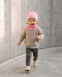 Dziewczynka w zestawie z uszami misia i kominem w kolorze Baby pink biegnie betonową ścieżką.