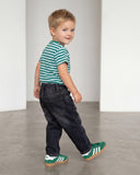Młody chłopak w koszulce PanPan 35 Bawełniany w paski dla chłopca - klasyczny krój na lato, ciemnych dżinsach i zielonych trampkach uśmiecha się, patrząc za siebie, gdy idzie po gładkiej podłodze w jasnym, minimalistycznym pokoju.
